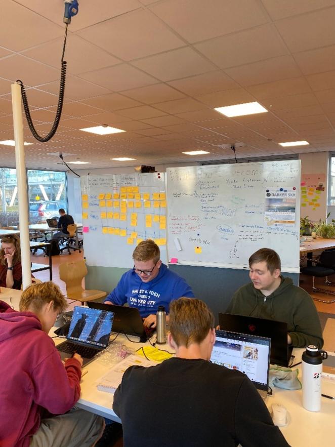 people looking at computer screens, and talking. On the background, a big whiteboard with many sticky notes